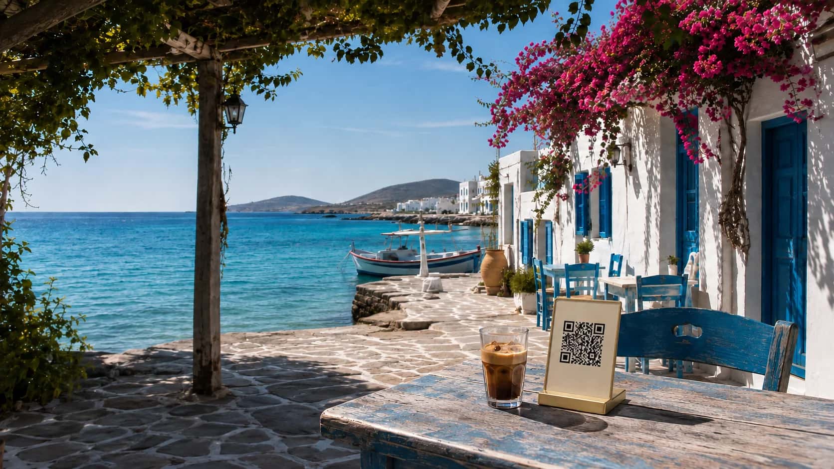 Ein griechisches Café am Meer mit blauem Himmel, türkisfarbenem Wasser und einem QR-Menüaufsteller auf einem Tisch.